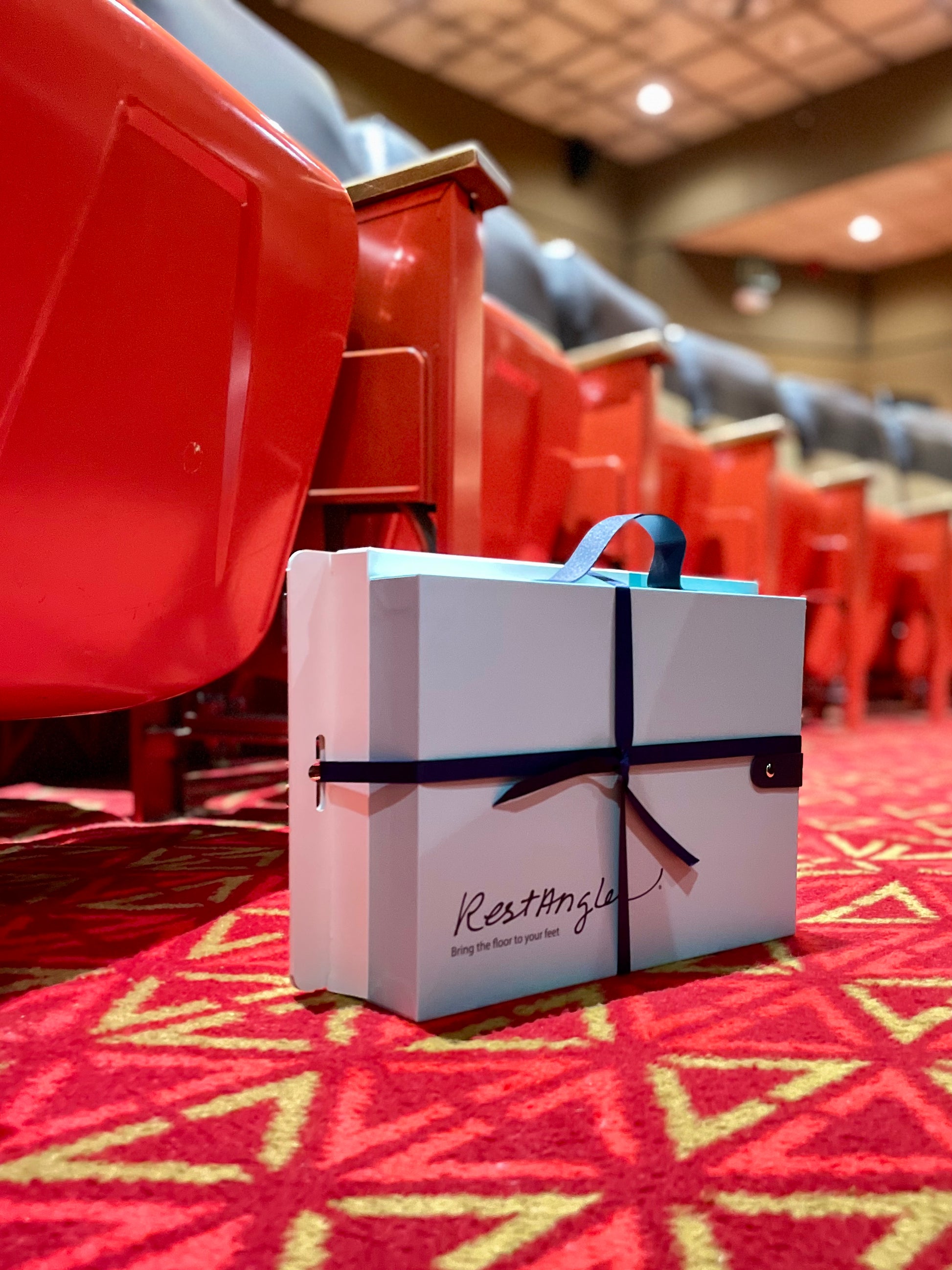 Light green gift box and white retail box packaged together with a dark blue bow on a red carpet in front of a row of movie theatre seats